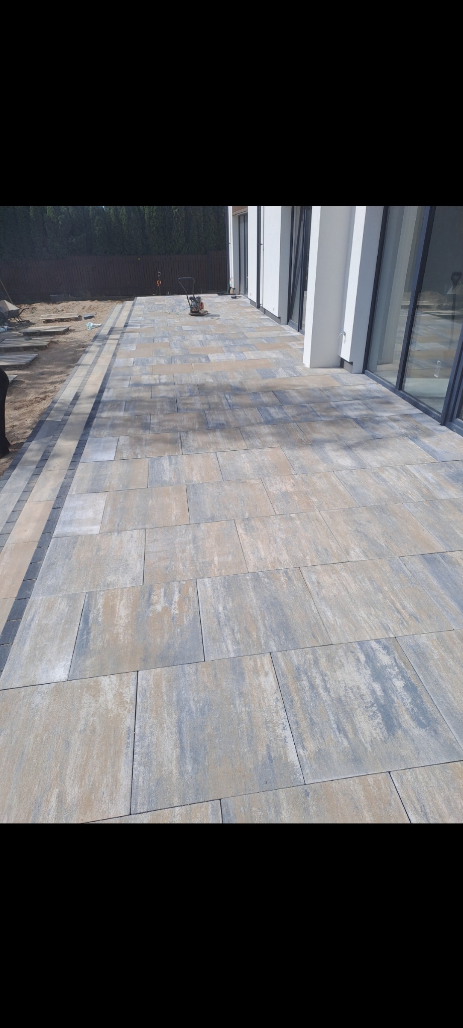 Nowoczesny taras z szaro-beżowej kostki brukowej o nieregularnym wzorze, obrzeża z ciemniejszej kostki, widoczna ubijarka wibracyjna, dom z białymi kolumnami w tle.