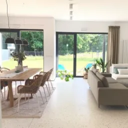 Jasne wnętrze domu z podłogą w drobny deseń, widok na ogród przez duże przesuwne okna, stół z wiklinowymi krzesłami i szary narożnik.