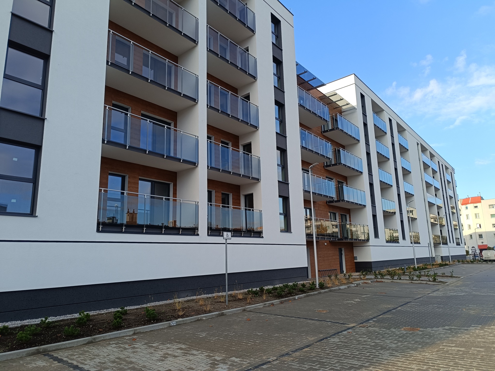 Nowoczesny budynek apartamentowy z balkonami zabezpieczonymi szkłem i metalowymi poręczami, widok z poziomu parkingu z kostki brukowej.