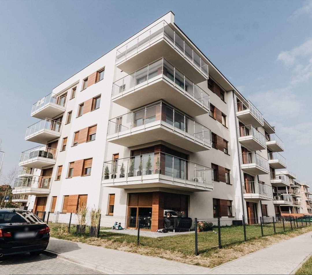 Nowoczesny budynek mieszkalny z balkonami zabezpieczonymi szkłem, widok z poziomu chodnika, z zaparkowanym samochodem w tle.