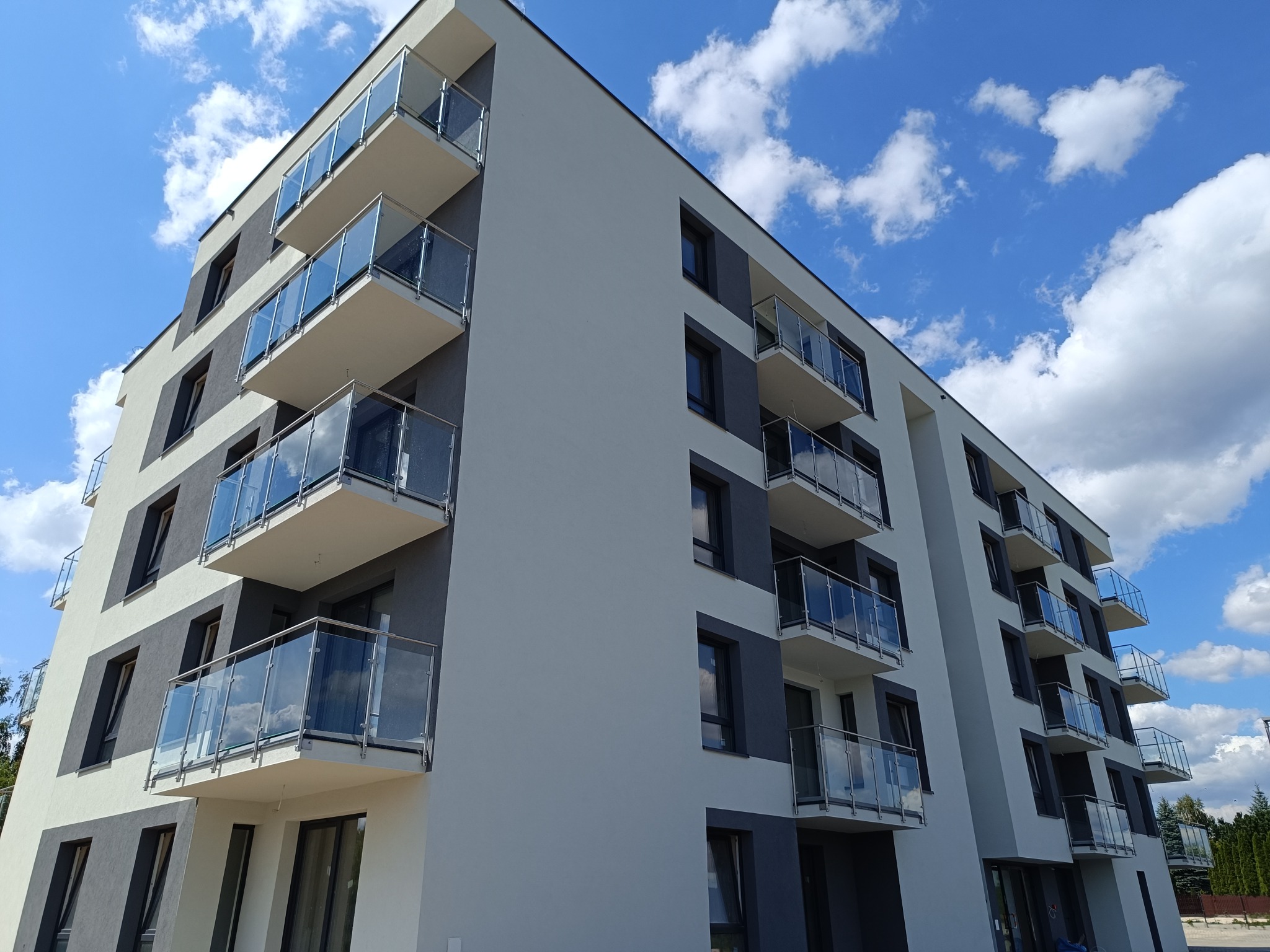 Nowoczesny budynek wielorodzinny z balkonami wyposażonymi w balustrady szklane z elementami ze stali nierdzewnej, widok z dołu na błękitne niebo z chmurami.