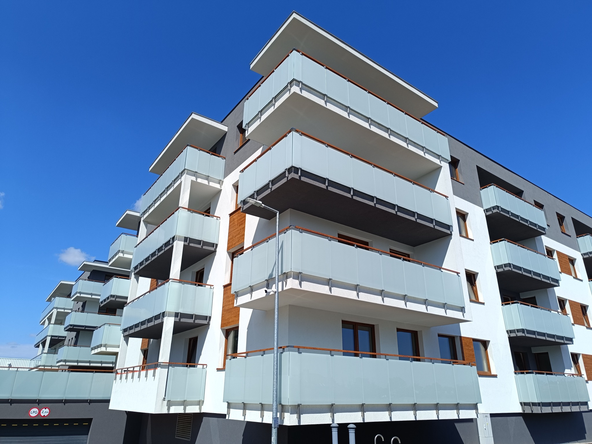 Nowoczesny budynek mieszkalny z balkonami zabezpieczonymi matowymi szklanymi panelami i drewnianymi poręczami, widok z dołu na tle błękitnego nieba.