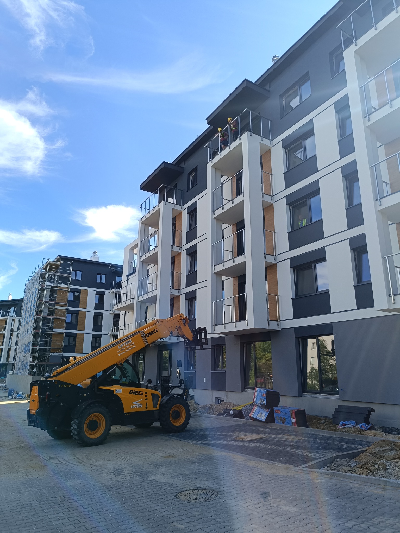 Montaż balustrad szklanych na balkonach nowego budynku wielorodzinnego za pomocą podnośnika teleskopowego Dieci, widoczni pracownicy w kaskach.