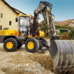 Żółto-czarna koparka kołowa z operatorem, podczas pracy na placu budowy, z widocznym wykopem i budynkami w tle, oświetlona naturalnym światłem słonecznym.