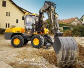 Żółto-czarna koparka kołowa z operatorem, podczas pracy na placu budowy, z widocznym wykopem i budynkami w tle, oświetlona naturalnym światłem słonecznym.