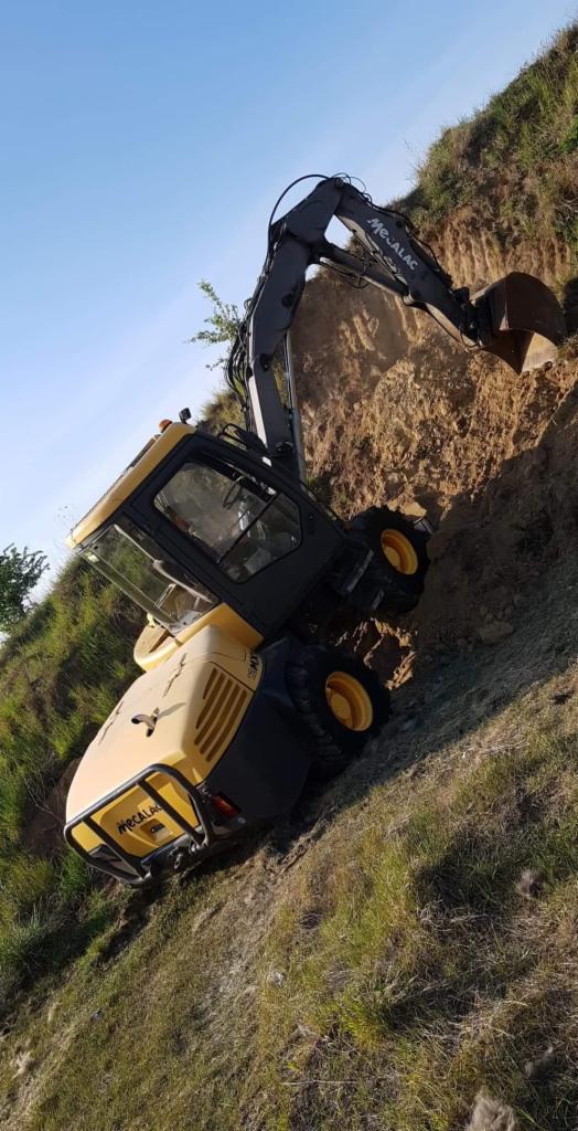 Żółto-szara koparka Mecalac na zboczu, z wysięgnikiem zagłębiającym się w ziemię, widok z dołu pod kątem.