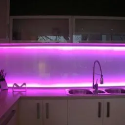 Kuchnia z podświetlanym panelem szklanym w kolorze fuksji, widoczny zlew z baterią i białe szafki.