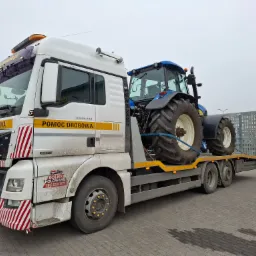 Laweta MAN TGX 26.440 z napisem 'Pomoc Drogowa' przewozi niebieski traktor New Holland na platformie. Na przedniej części lawety widoczne logo firmy 'Dolny Holowanie' oraz czerwono-białe pasy.