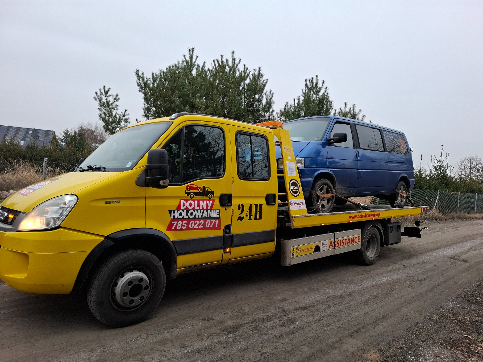 Żółty pojazd pomocy drogowej z niebieskim vanem na lawecie, widoczne logo firmy 'DOLNY HOLOWANIE' i numer telefonu, tło stanowią drzewa i pochmurne niebo.