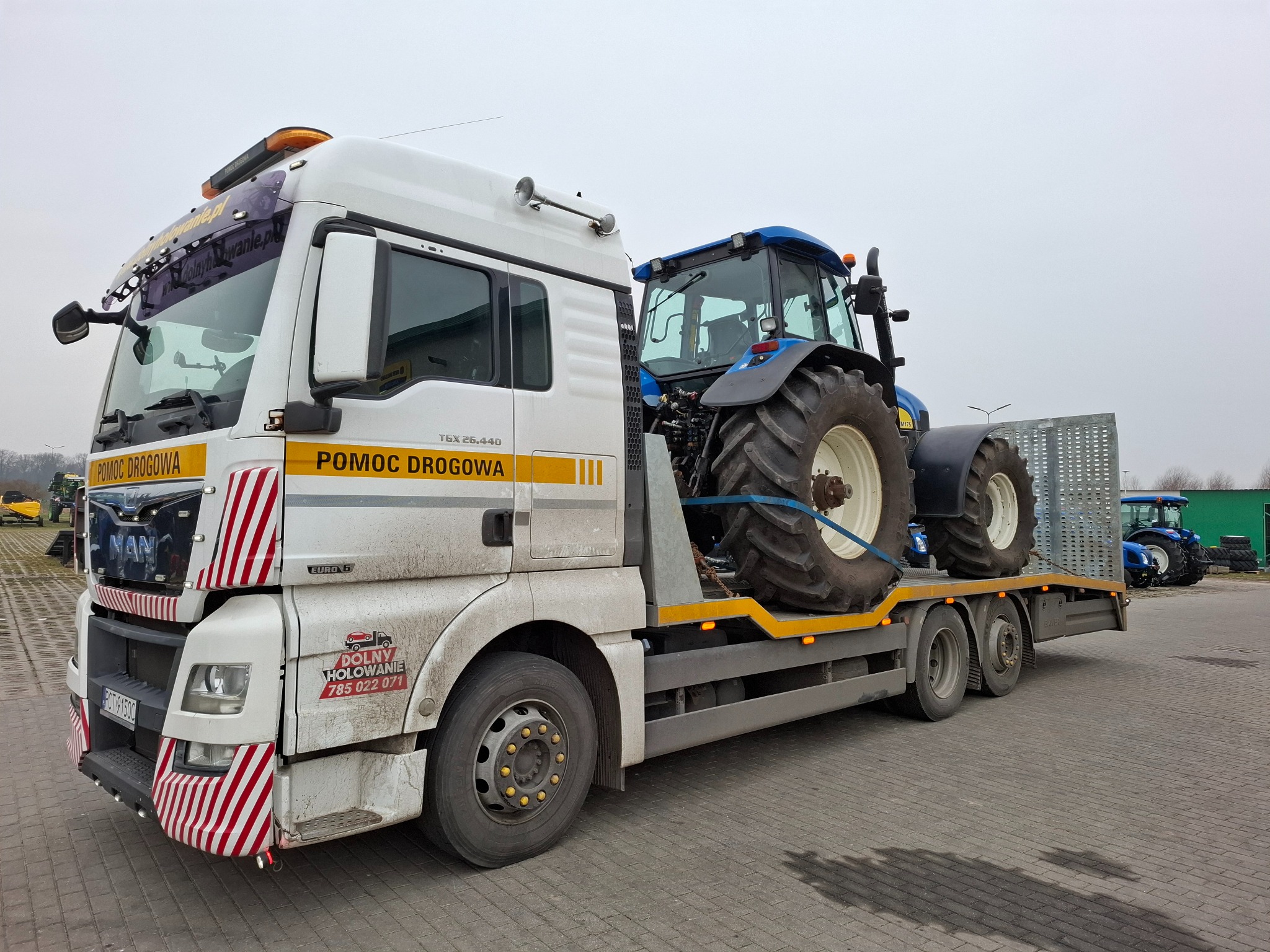 Laweta MAN TGX 26.440 z napisem 'Pomoc Drogowa' przewozi niebieski traktor New Holland na platformie. Na przedniej części lawety widoczne logo firmy 'Dolny Holowanie' oraz czerwono-białe pasy.