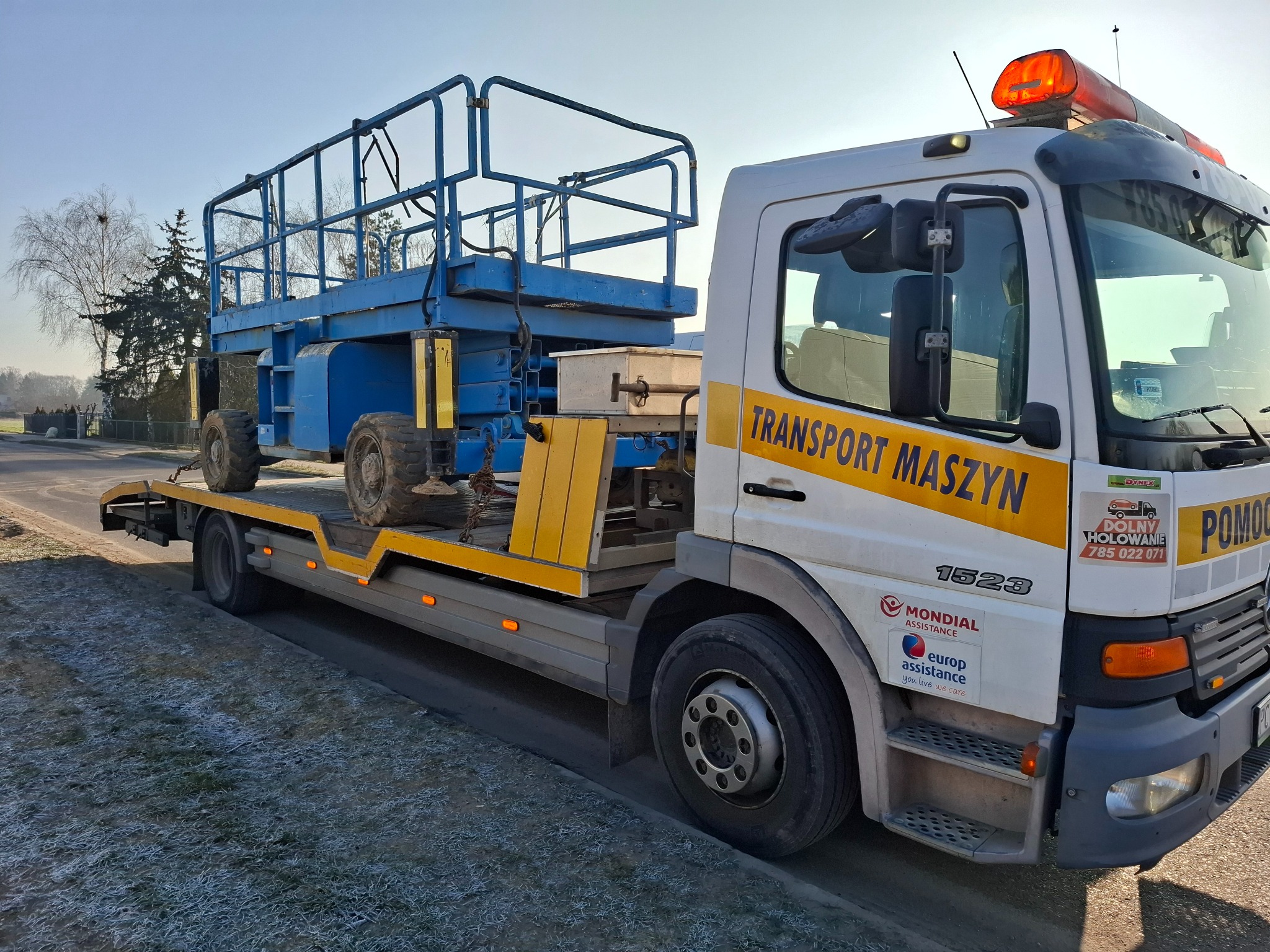 Ciężarówka z platformą załadunkową przewożąca podnośnik koszowy w słoneczny dzień, widoczne logo firmy transportowej i numer telefonu.