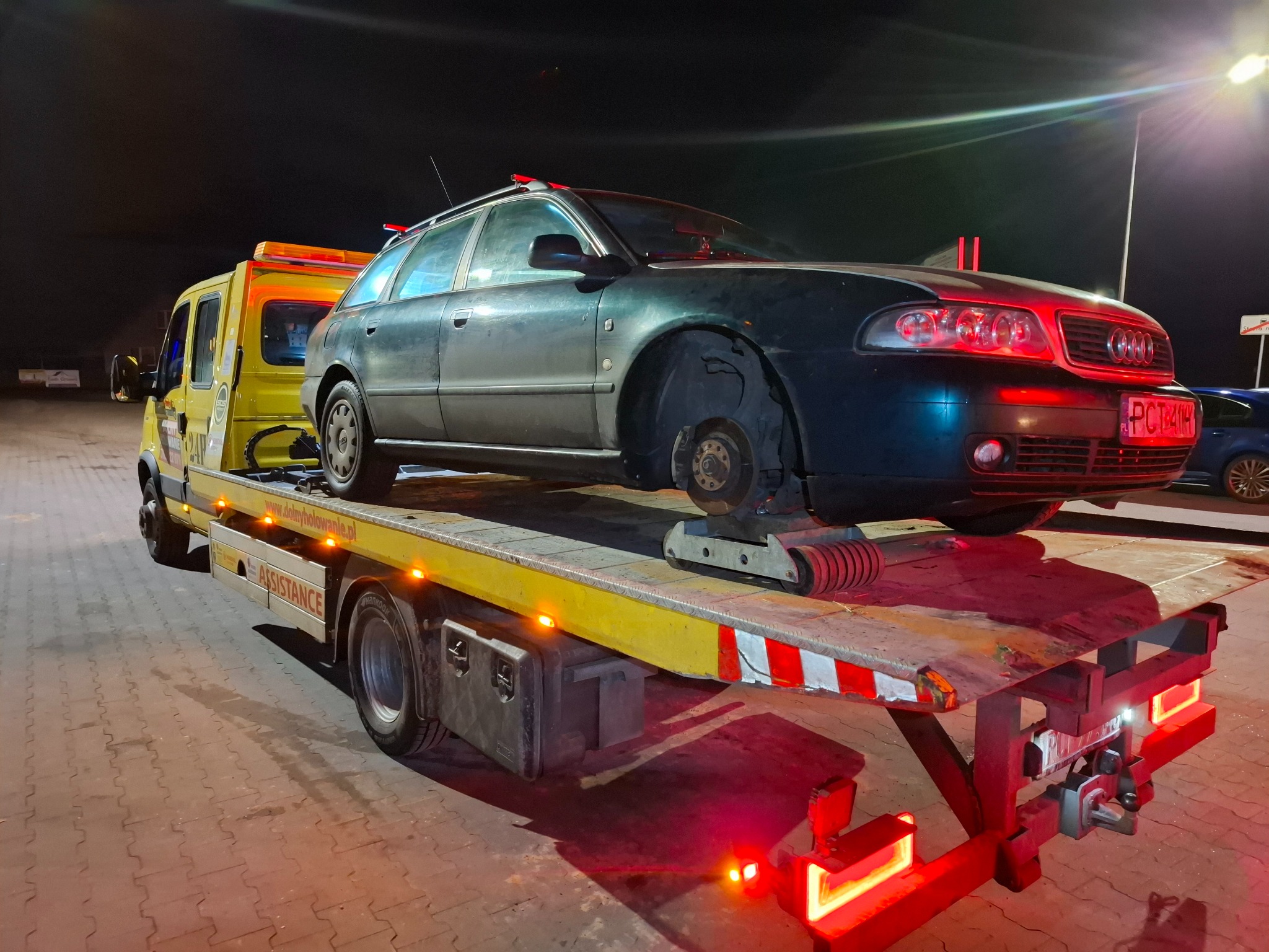 Uszkodzony ciemny samochód osobowy marki Audi bez koła, zabezpieczony na żółtej platformie pojazdu pomocy drogowej z włączonym oświetleniem ostrzegawczym, nocą na parkingu.