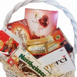 Biały wiklinowy kosz wypełniony słodyczami, kawą Lavazza i herbatą Warztee, zapakowany w ozdobny papierowy wypełniacz.