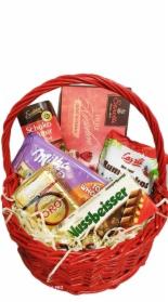 Czerwony wiklinowy kosz wypełniony różnorodnymi słodyczami i przekąskami, w tym czekoladami w opakowaniach w odcieniach brązu, fioletu i czerwieni, ułożonymi na jasnym, papierowym wypełniaczu.