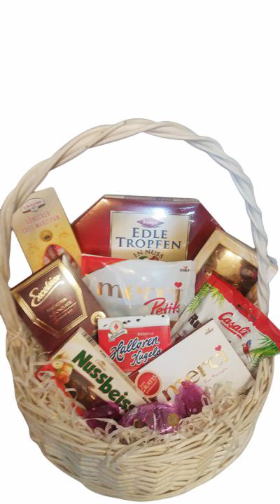 Wiklinowy kosz wypełniony różnorodnymi słodyczami, w tym czekoladkami Merci, Nussbeisser i Edle Tropfen, zapakowanymi w ozdobne opakowania.