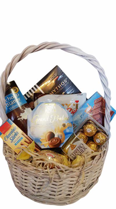 Wypełniony po brzegi wiklinowy kosz z różnorodnymi słodyczami: czekoladami, pralinami, batonikami, w tym marki Ferrero Rocher, Monk Rotti i Mythos, na białym tle.