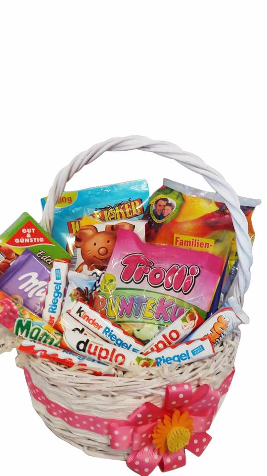 Biały wiklinowy koszyk wypełniony różnorodnymi słodyczami, w tym batonami Kinder Riegel Duplo, żelkami Trolli, cukierkami i przekąskami, ozdobiony różową wstążką w białe kropki i żółtym kwiatem.
