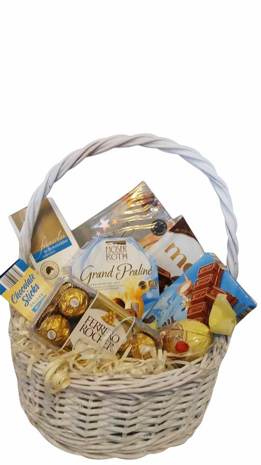 Biały wiklinowy kosz wypełniony różnorodnymi słodyczami, w tym czekoladkami Ferrero Rocher, Moser Roth Grand Praline i czekoladowymi batonikami, owinięty wstążką.
