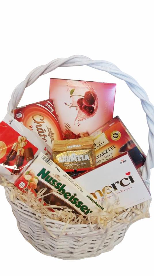 Biały wiklinowy kosz wypełniony słodyczami, kawą Lavazza i herbatą Warztee, zapakowany w ozdobny papierowy wypełniacz.