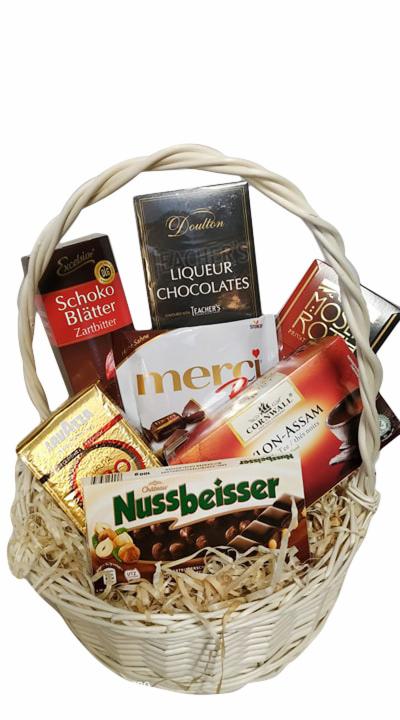 Biały, wiklinowy kosz wypełniony różnorodnymi markami czekolad, w tym Nussbeisser, Merci, Doulton Liqueur Chocolates i Schoko Blätter Zartbitter.