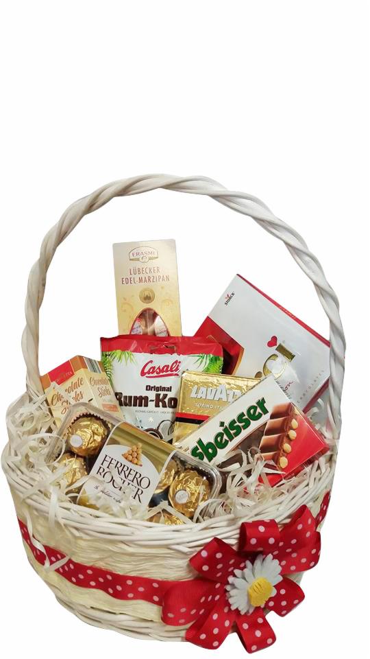 Biały wiklinowy kosz wypełniony różnorodnymi słodyczami, w tym czekoladkami Ferrero Rocher, marcepanem Lubecker, batonami czekoladowymi i pralinami, ozdobiony czerwoną wstążką w białe kropki...