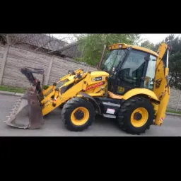 Żółta koparko-ładowarka JCB 3CX z łyżką z przodu, zaparkowana na asfaltowej drodze, w tle drewniany płot i drzewa.