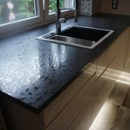 Czarny, teksturowany blat kuchenny z metalowym zlewem i baterią, oświetlony światłem z okna i ledowym oświetleniem szafek, z widocznymi płytkami ceramicznymi w tle.