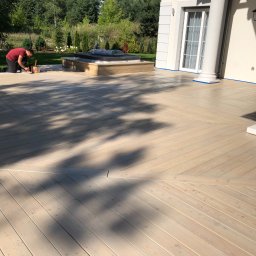 Tomasz  Simm - Malowanie drewnianego tarasu w Warszawie: mężczyzna klęczy i maluje deski jasną farbą, widoczny fragment domu z kolumnami i zieleń w tle.