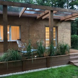 Tomasz  Simm - Drewniana konstrukcja z zadaszeniem z przezroczystego tworzywa, połączona z budynkiem, z roślinami w drewnianych donicach, oświetlona wieczornym światłem, z widocznymi schodami wejściowymi.