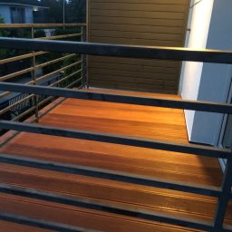 Tomasz  Simm - Nowoczesny balkon z drewnianą podłogą i metalową balustradą, widok z góry, ciepłe oświetlenie podkreśla fakturę drewna. Minimalistyczny design.
