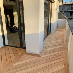 Tomasz  Simm - Nowoczesny taras drewniany z jasnych desek, ułożonych pod kątem, z widocznym fragmentem balkonu i elementami wykończeniowymi. W tle widoczne narzędzia pracy.