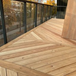 Tomasz  Simm - Nowoczesny balkon z drewnianą podłogą ułożoną we wzór jodełki, szklana balustrada z widokiem na jesienne drzewa. Naturalne oświetlenie podkreśla ciepły odcień drewna.