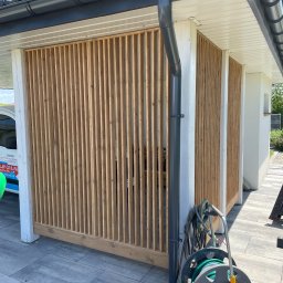 Tomasz  Simm - Nowoczesna, drewniana ażurowa ściana osłonowa przy budynku z szarą rynną. Widoczny fragment kostki brukowej i zwinięty wąż ogrodowy.