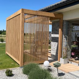 Tomasz  Simm - Nowoczesna, drewniana konstrukcja z pionowych listew, przylegająca do domu z kamienną elewacją. Oświetlona kula i rośliny na żwirowym podłożu.