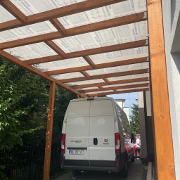 Tomasz  Simm - Drewniana konstrukcja z przezroczystym zadaszeniem, prawdopodobnie wiata garażowa, z zaparkowanym białym busem. Widok od dołu, z perspektywy parkingu.