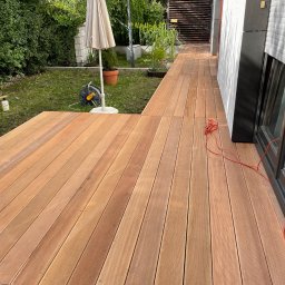 Tomasz  Simm - Nowoczesny taras z desek kompozytowych w odcieniach brązu, przylegający do budynku z jasną elewacją. Widoczny parasol ogrodowy i zadbana zieleń w tle.