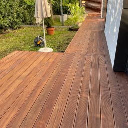 Tomasz  Simm - Nowoczesny taras z desek kompozytowych w ogrodzie, z widocznym parasolem i roślinami w donicach. Czyste linie, ciepłe oświetlenie, zadbana przestrzeń.