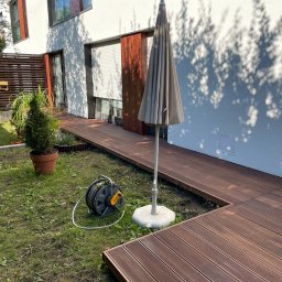 Tomasz  Simm - Nowoczesny taras z desek kompozytowych w ogrodzie, z widocznym zwiniętym wężem ogrodowym i parasolem. Cień drzew na białej ścianie domu.