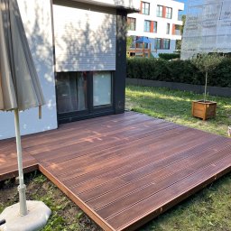 Tomasz  Simm - Nowoczesny taras z brązowych desek kompozytowych przylegający do budynku z roletami, z parasolem i widokiem na budynek w rusztowaniach.