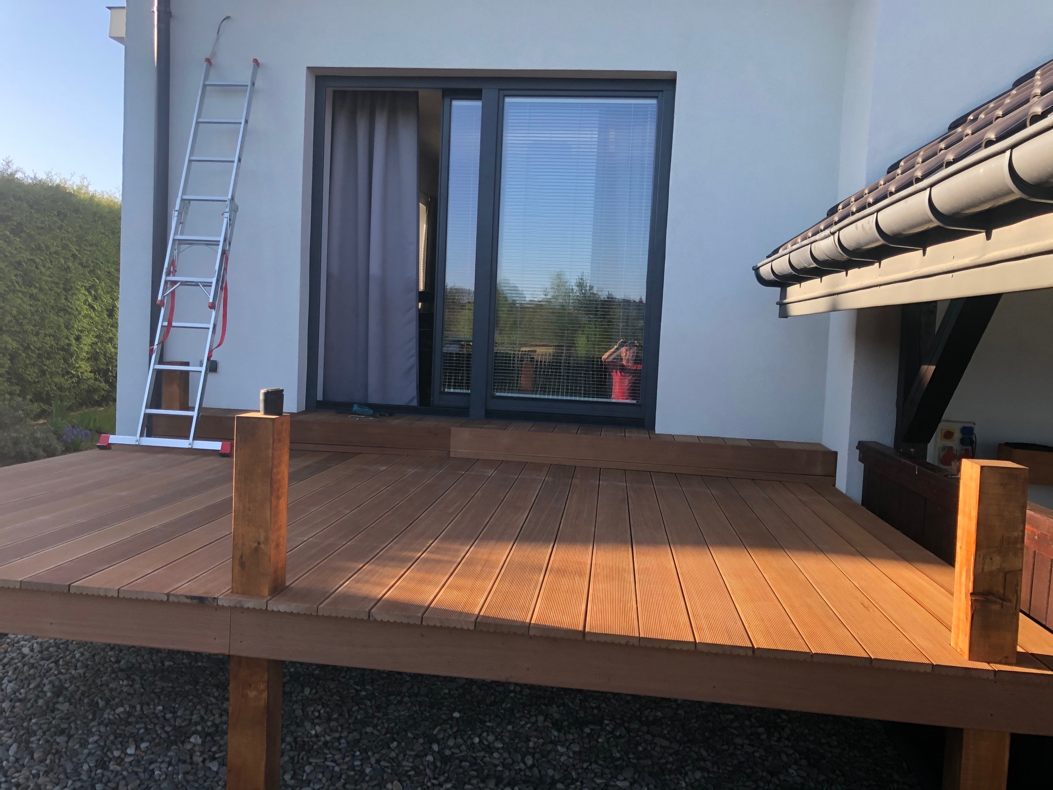 Nowoczesny taras z desek kompozytowych w trakcie budowy, widoczna drabina oparta o ścianę domu i osoba robiąca zdjęcie w odbiciu szyby.