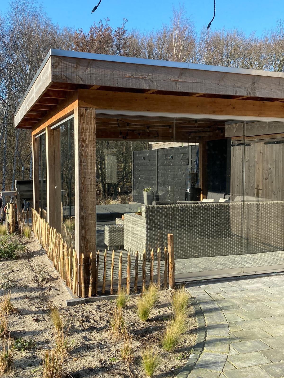 Zadaszony taras z drewnianą konstrukcją, szklanymi ścianami, wiklinowymi meblami wypoczynkowymi i niskim płotkiem z drewnianych kołków.