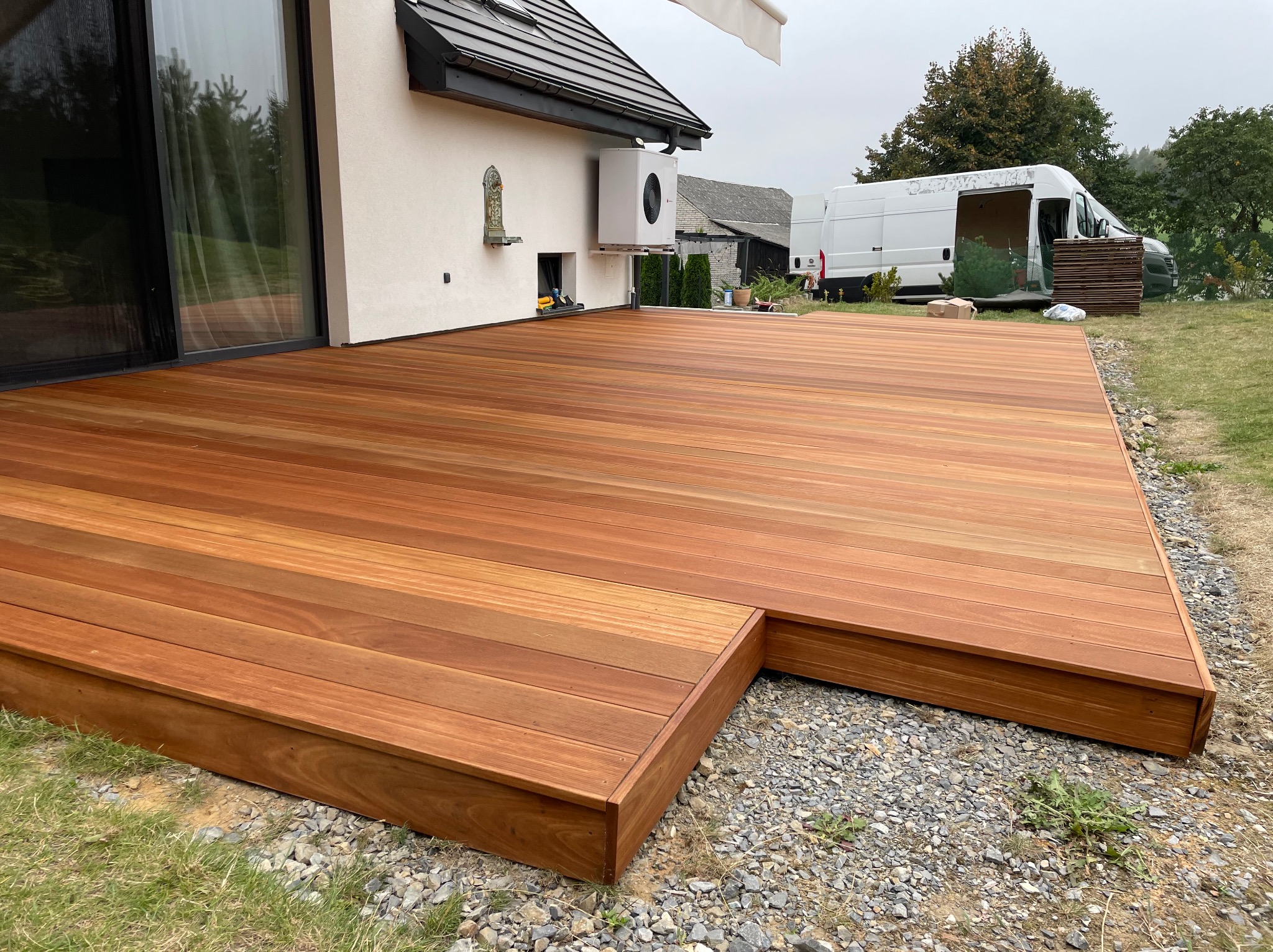 Nowoczesny taras z drewna egzotycznego, z niskim stopniem, na żwirowym podłożu. W tle biały budynek z klimatyzatorem i otwarty bus dostawczy z deskami.