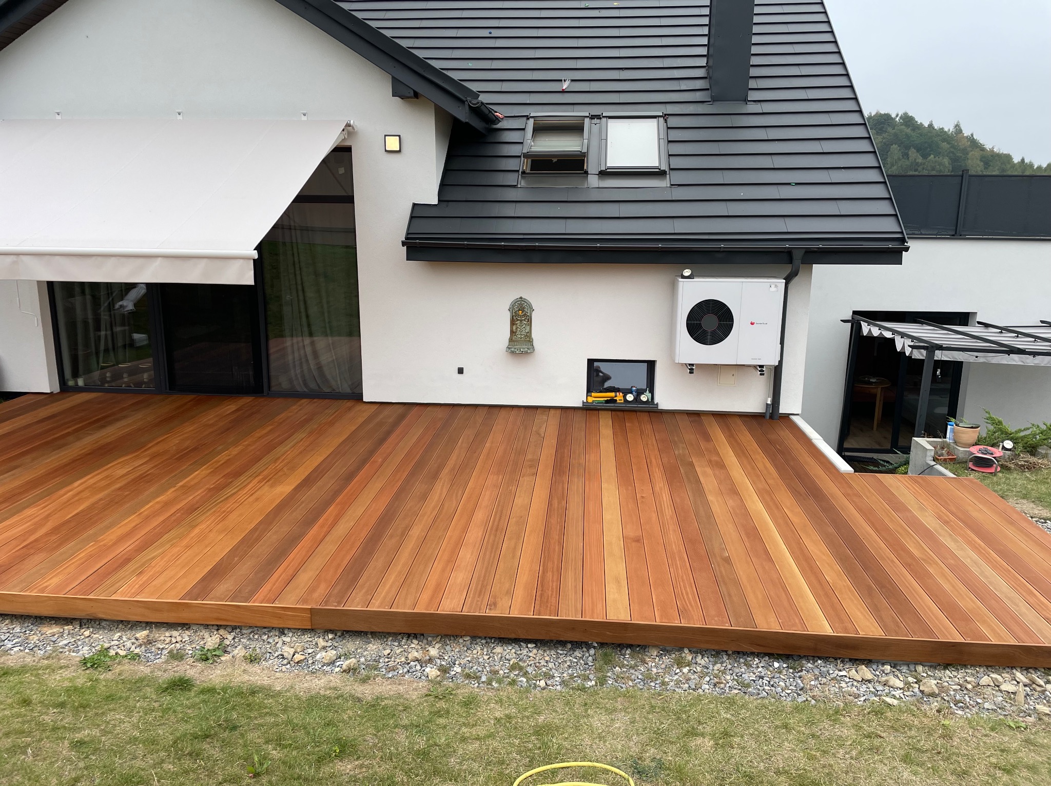 Nowoczesny taras z desek w ciepłym odcieniu drewna, przylegający do białego domu w Warszawie. Widoczna markiza i klimatyzator na elewacji.