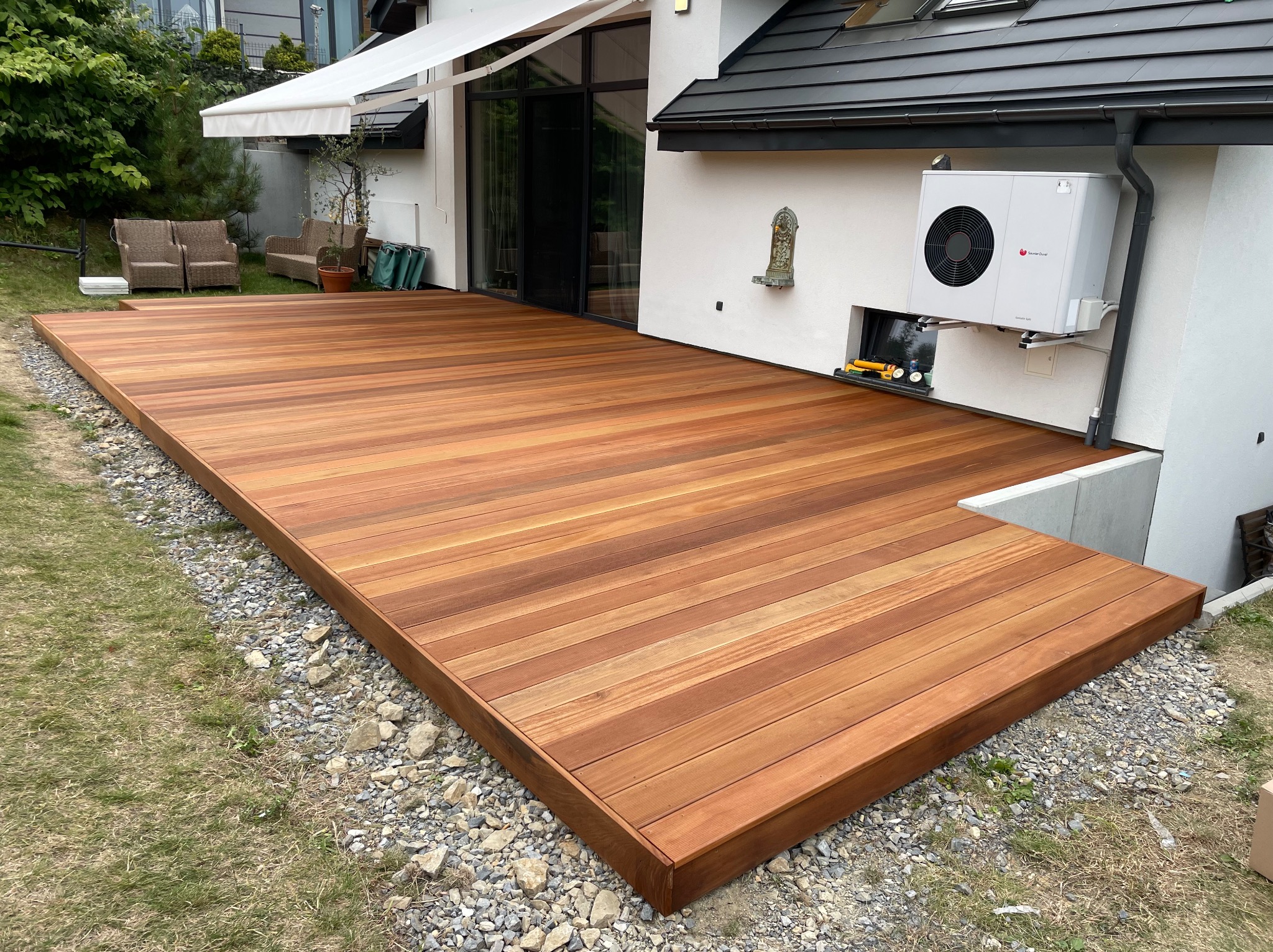 Rozległy, drewniany taras z desek w ciepłym odcieniu brązu, przylegający do nowoczesnego domu z białymi ścianami i szarym dachem. Kamieniste podłoże i zieleń w tle.