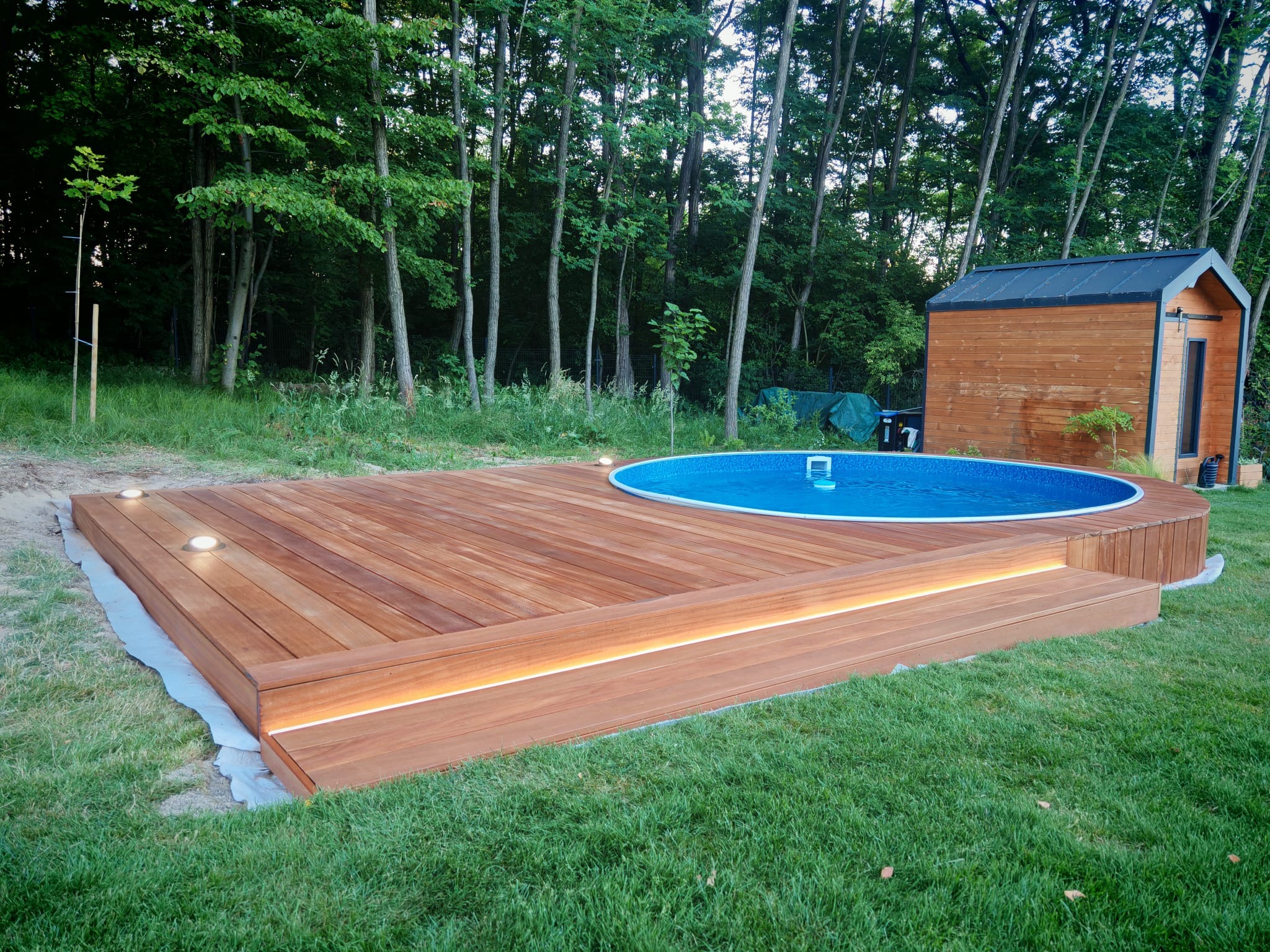 Drewniany taras z okrągłym basenem i oświetleniem wbudowanym w deski, otoczony zielenią i drzewami. Obok stoi mała drewniana budka.