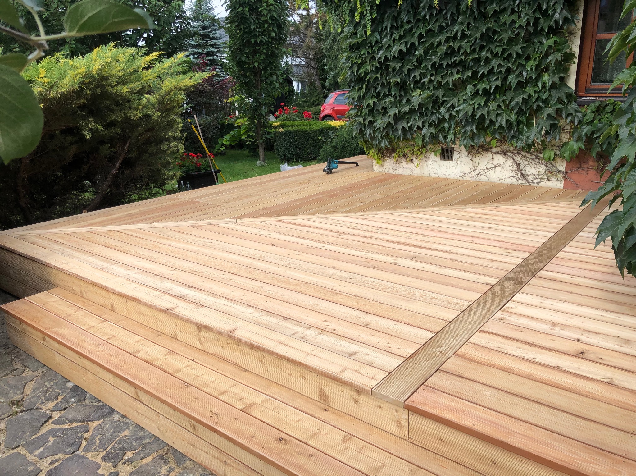 Nowoczesny taras drewniany z schodami, wykonany z jasnego drewna, otoczony zielenią. Na tarasie leży piła akumulatorowa. Widok na ogród z czerwoną roślinnością.