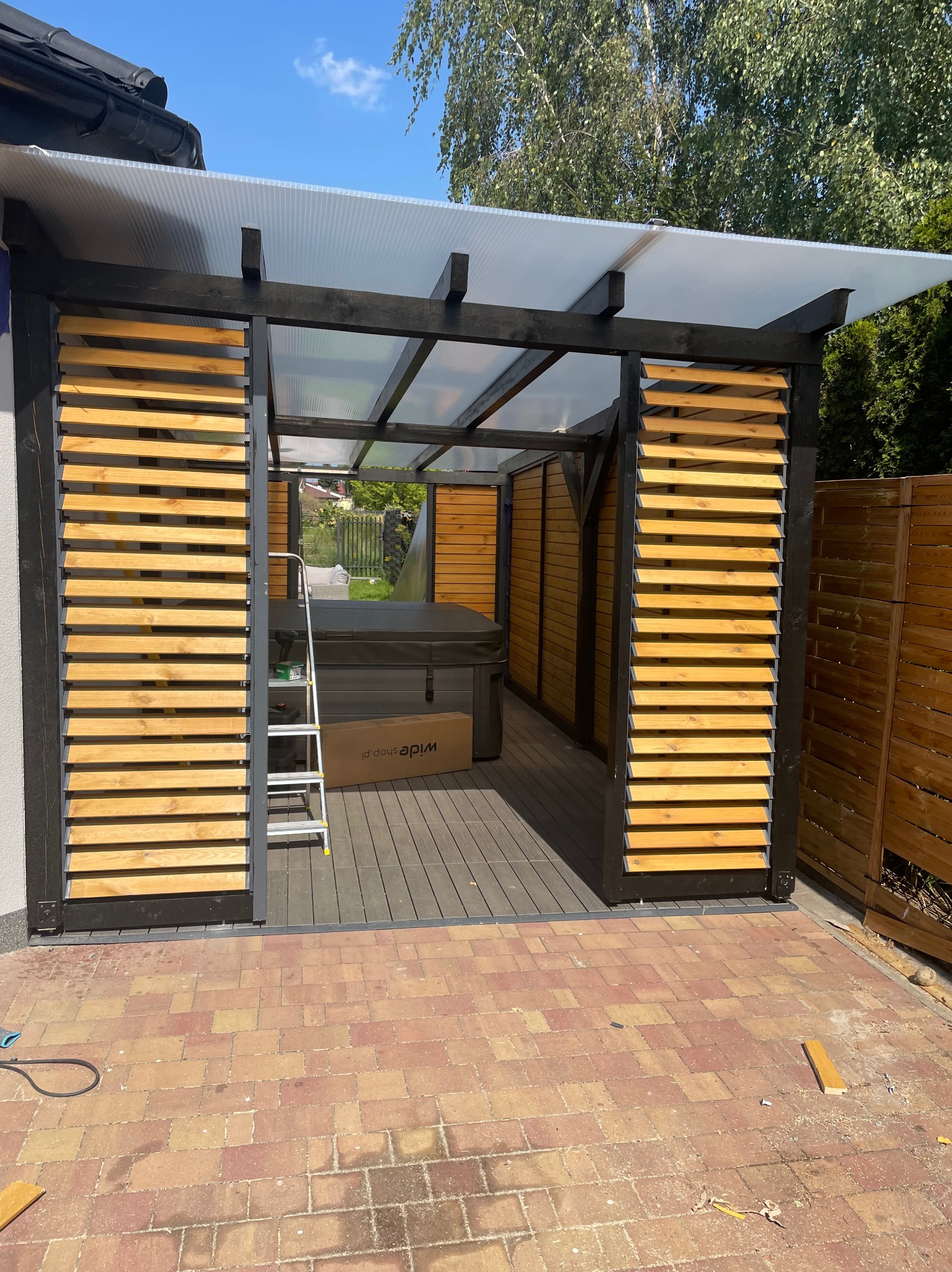 Nowoczesna, czarna konstrukcja z drewnianymi żaluzjami i przezroczystym zadaszeniem. Wewnątrz jacuzzi, obok drabina i karton. Na podłożu kostka brukowa.