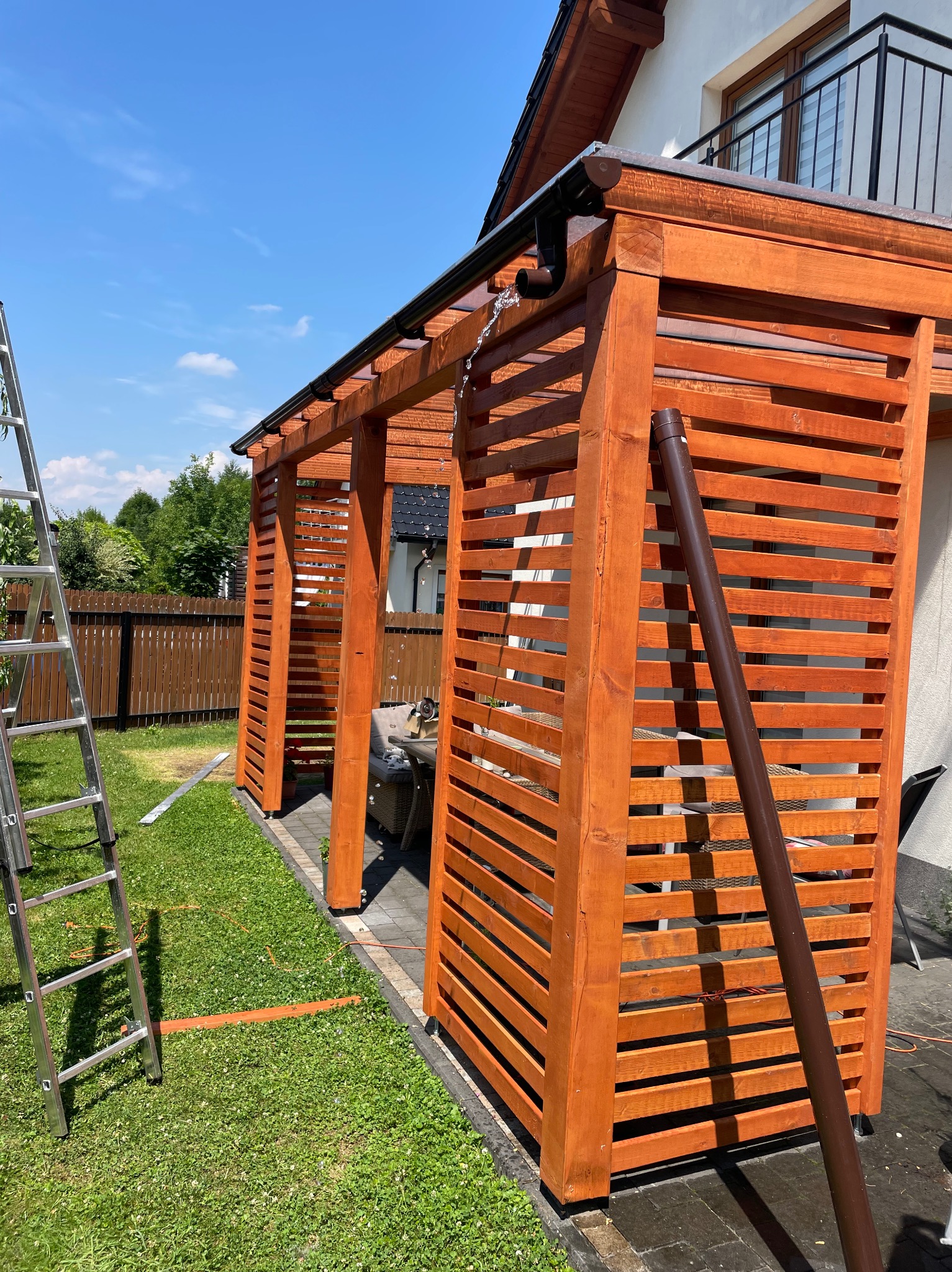 Brązowa, drewniana pergola z lamelami w ogrodzie, z widoczną drabiną i zlewką wody z rynny. Wyraźne słońce i cień. Detal konstrukcji.