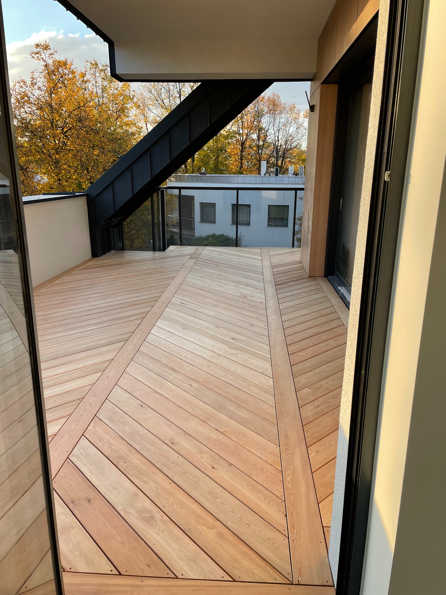 Nowoczesny taras z jasnego drewna ułożonego we wzór jodełki, widok z wnętrza, jesienne drzewa w tle. Minimalistyczna architektura.