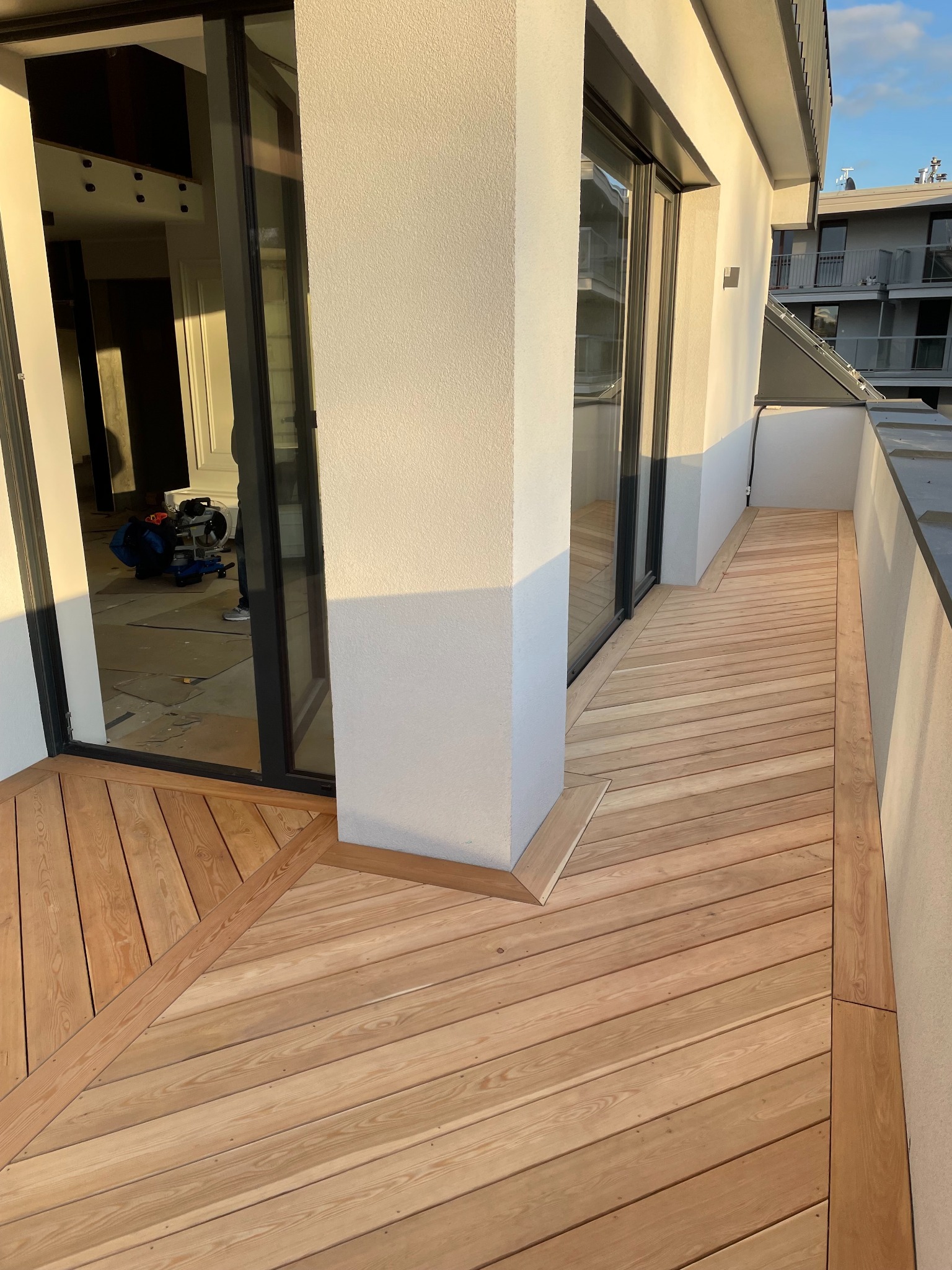 Nowoczesny taras drewniany z jasnych desek, ułożonych pod kątem, z widocznym fragmentem balkonu i elementami wykończeniowymi. W tle widoczne narzędzia pracy.